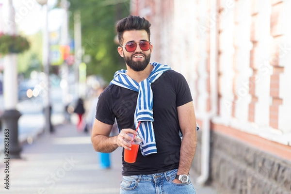 Fototapeta Handsome cheerful young arabian student with a stylish mustache and a beard with juice in his hands, walking around the city after a working day. Concept of positive and rest.