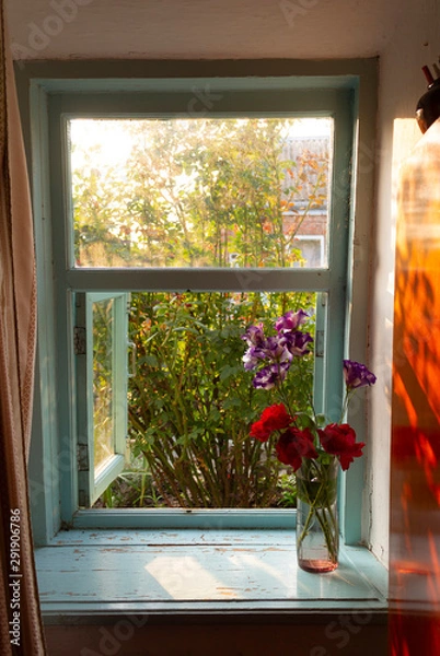 Fototapeta The window in the old residential building is open in the morning in the summer, the rose outside the window and a bouquet of flowers on the windowsill, the morning sun sets in the room.