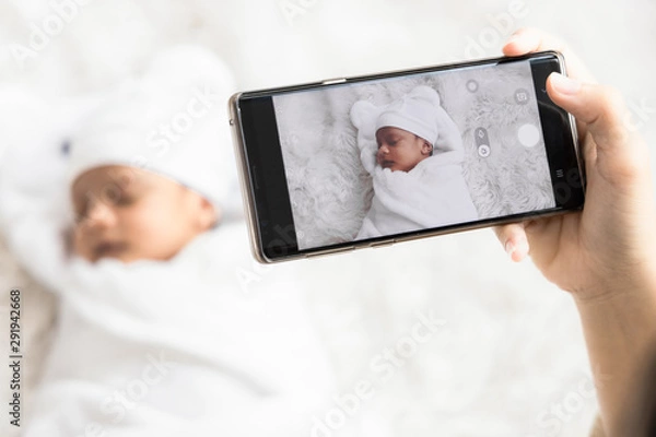 Fototapeta Mother 's hand holding smartphone use to taking photo cute newborn baby for memories and pose in social network.