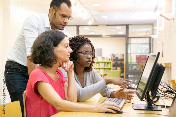 Fototapeta Tutor helping students in computer class. Man and women sitting and standing at desk, using desktop, pointing at monitor and talking. Education concept