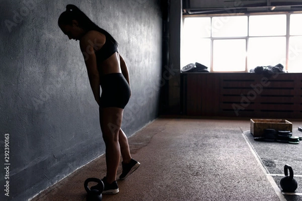 Obraz Fit anonymous woman prepare for training with weights - kettlebells. Light from a window. Silhouette of athlete, fitness model.