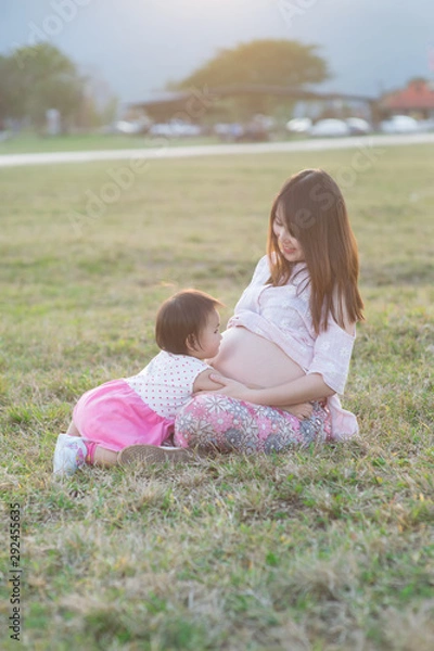 Obraz baby girl  hugging asian beautiful pregnant woman on grass field in evening light with sunset and mountain background, Good health and relax concept, family relationship.