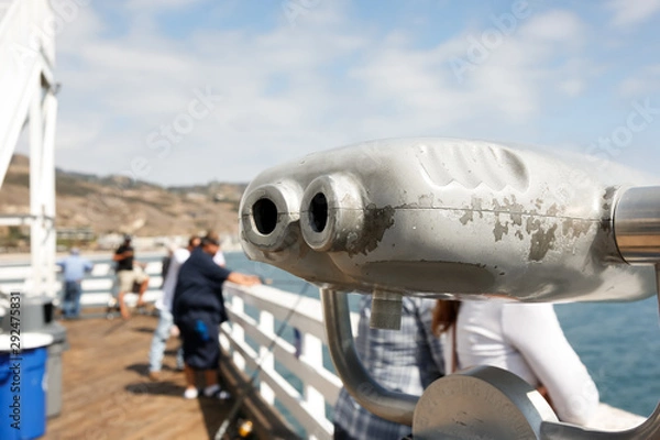 Fototapeta A closeup view of pier binoculars, featuring people in the background fishing and enjoying the view.