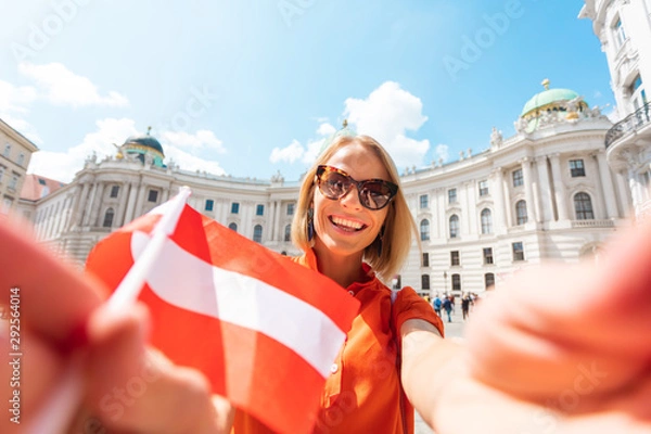 Obraz Young happy woman tourist with the flag of Austria makes selfie photo on the background of Michaelplatz and Hofburg in Vienna, Austria