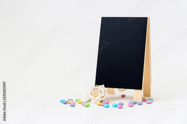 Fototapeta Blank black chalkboard easel with pastel-colored hard-shelled candies on a white background