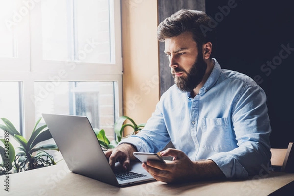 Fototapeta Creative worker using digital devices and programs in project. Corporate executive sitting at office desk, typing on laptop. Manager making important business phone calls on smartphone, checking email