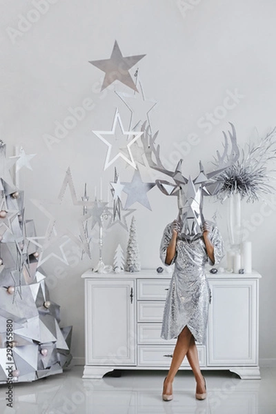Fototapeta Beautiful and modish young woman with silver bright makeup, in bright silver dress and in a silver mask of deer posing near the alternative silver Christmas tree in the interior