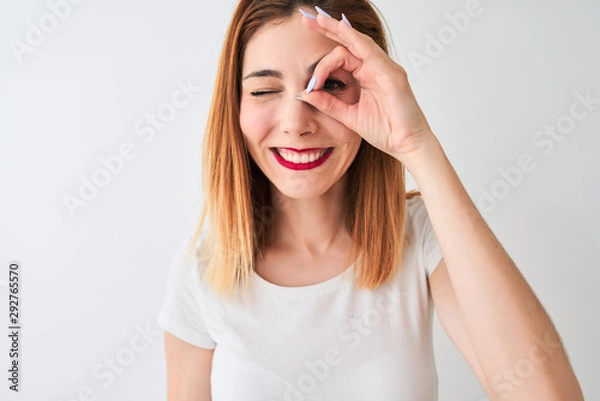 Fototapeta Beautiful redhead woman wearing casual t-shirt standing over isolated white background with happy face smiling doing ok sign with hand on eye looking through fingers