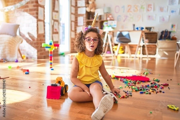 Fototapeta Beautiful toddler wearing glasses and unicorn diadem sitting on the floor at kindergarten afraid and shocked with surprise expression, fear and excited face.