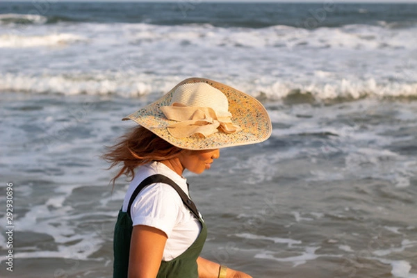 Fototapeta A young African lady in a sun hat playing by the beach