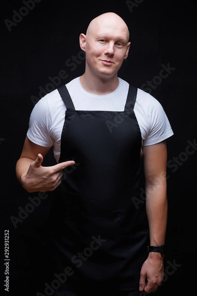 Obraz Strong handsome smiling bald guy wearing black apron pointing his finger on his chest. Butcher, baker, chef or waiter concept. Mockup for restaurants, grossery shops, bakery, butchery.