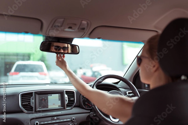 Fototapeta girl in passenger compartment of car looks into bright light of rear view, checks children in back row of seats, parking shopping center, reversing car. Woman in summer sunglasses in city.