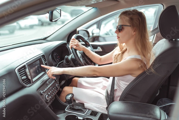 Fototapeta girl in car selects application on touch screen, navigation application on Internet, strapped holds wheel, left-hand traffic, wheel on right side. Woman in summer sunglasses in city.