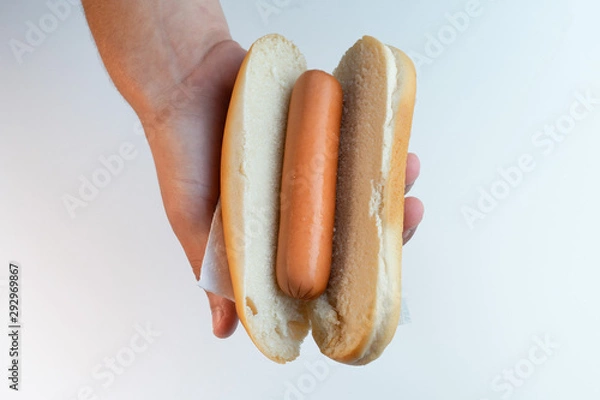 Fototapeta Hand holding a Hot dog bun with sausage on a white background. Sandwich in the palm of hand. Business lunch. Top view.