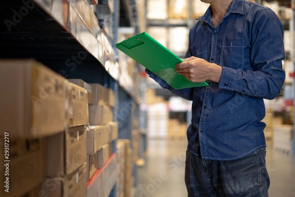 Fototapeta Manager man worker doing stocktaking of product management in cardboard box on shelves in warehouse. Physical inventory count.. Male professional assistant checking stock in factory.