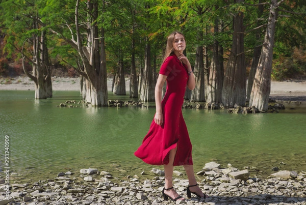 Fototapeta a beautiful, young brown-haired woman in a red dress on the background of a morning lake with cypresses, a girl with a slender figure posing against nature
