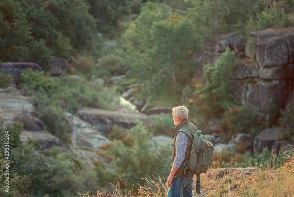 Fototapeta Back view of tourist man with a gray beard with a backpack on his shoulders against the backdrop of the gorge, rocks and stones, the concept of tourism and outdoor activities in old age