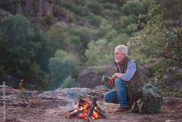 Fototapeta Eldery tourist with gray hair and beard sits on the edge of a canyon near a burning fire with a mug in his hands, against the backdrop of the sun, mountains and trees