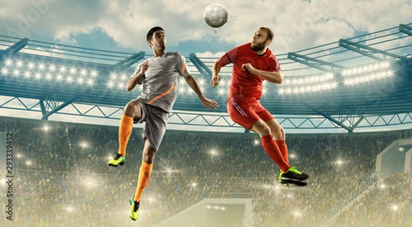 Obraz Soccer players fight for a ball in air. Jump shot. Soccer field with tribunes