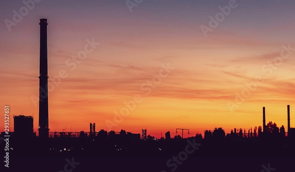 Fototapeta Sunset in the industrial zone. A factory with a tall chimney and glowing towers. Beautiful sky with clouds.