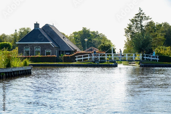 Fototapeta Beautiful scenery during summer with a small bridge and cycling tourists and with a typical dutch house next to the canal in Ossenzijl, National Park Weerribben-Wieden, Overijssel, The Netherlands.