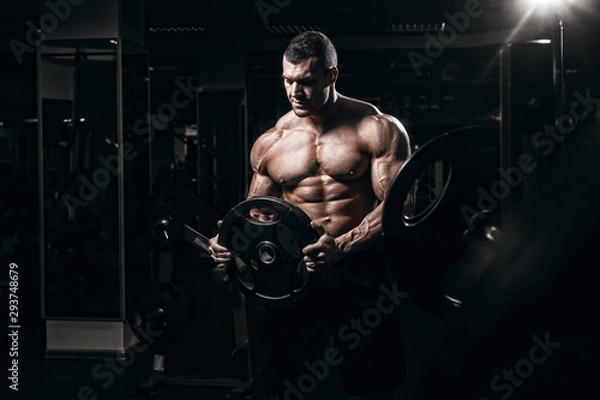Fototapeta Muscular man bodybuilder training in gym and posing. Fit muscle guy workout with weights and barbell