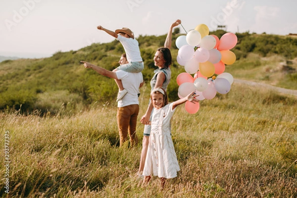 Fototapeta Beautiful family of four taking a walk in the green mountain fields carrying colourful baloons smiling and enjoying their holiday .