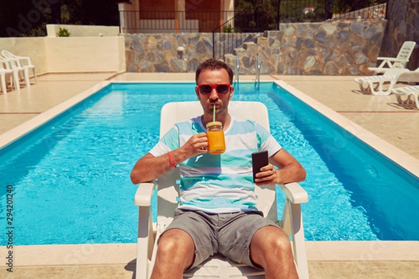 Fototapeta Attractive man using cellphone while drinking juice near swimming pool.