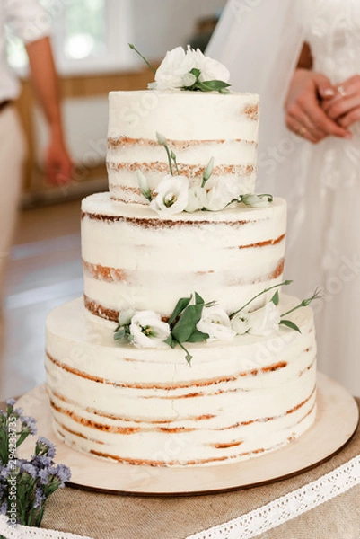 Fototapeta Wedding ceremony. Beautiful pie on the table. Elegant wedding cake decorated with white eustoma and vanilla glaze with bride and groom on background. Brides cut cake together. Close up
