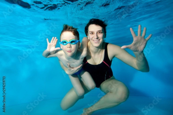 Obraz A little boy and his smiling mother swim and pose underwater in the pool. They hug and look at the camera. A child with swimming glasses. Portrait. Family entertainment. Underwater photography