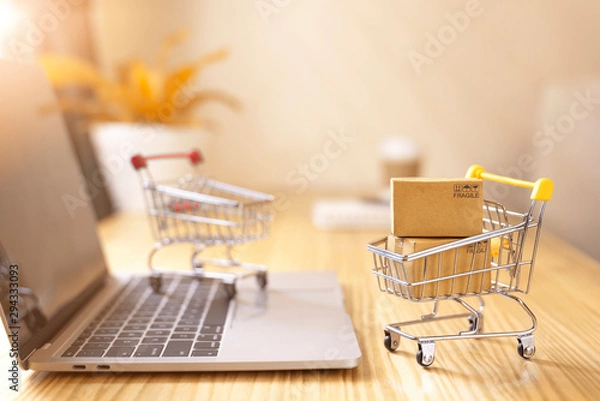 Fototapeta Online shopping and delivery service concept.Brown paper boxs in a shopping cart with laptop keyboard on wood table in office background.Easy shopping with finger tips for consumers.