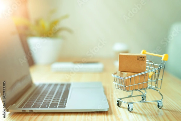 Fototapeta Brown paper boxs in a shopping cart with laptop keyboard on wood table in office background.Easy shopping with finger tips for consumers.Online shopping and delivery service concept.