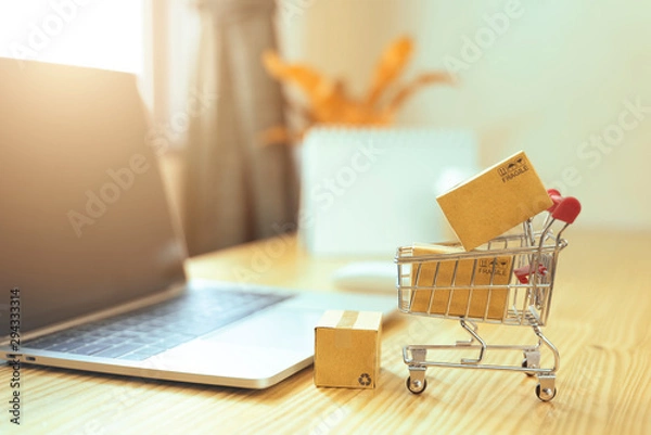 Fototapeta Brown paper boxs in a shopping cart with laptop keyboard on wood table in office background.Easy shopping with finger tips for consumers.Online shopping and delivery service concept.