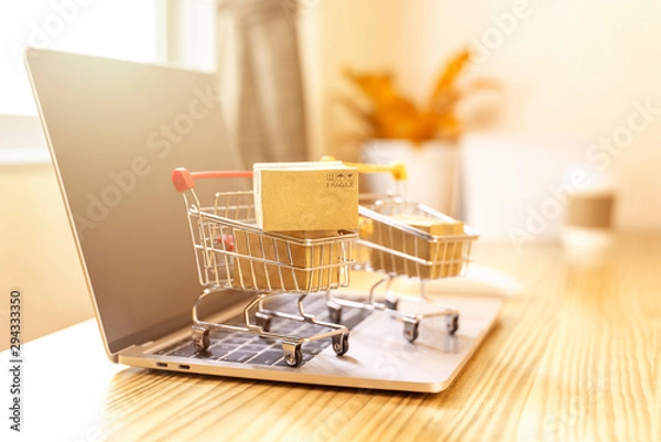 Fototapeta Brown paper boxs in a shopping cart with laptop keyboard on wood table in office background.Easy shopping with finger tips for consumers.Online shopping and delivery service concept.