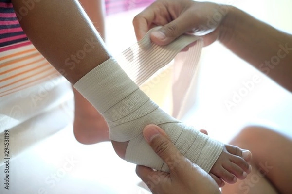 Obraz Injured kid ankle. Father rewinds kid’s leg with an elastic ankle support.