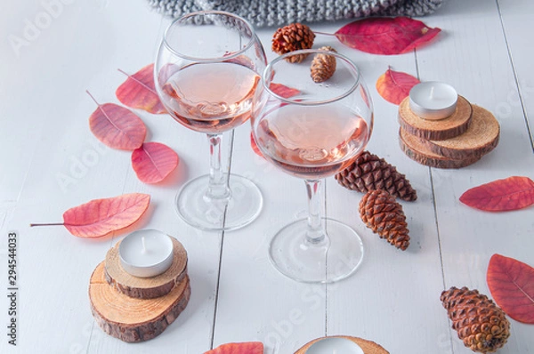 Fototapeta Small candles, two glasses with rose wine, cones, dry red leaves, gray scarf knitted on a white wooden table. Hello, Autumn. Cozy autumn background.