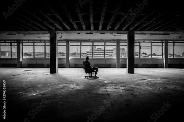 Obraz Man sitting in front of a large window in an empty hall of an abandoned building - black and white