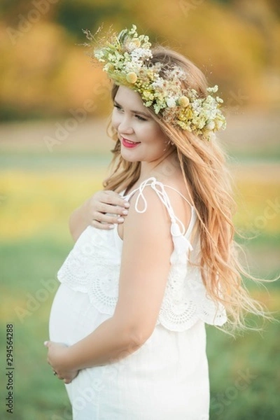 Fototapeta Portrait of a pregnant woman in the sun. Young beautiful pregnant woman with a wreath on her head in the field. Motherhood. Warm autumn.