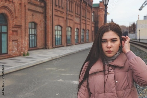 Obraz A young pretty pregnant girl in a pink warm down jacket walks near the old railway station. Late autumn, the cold is coming.