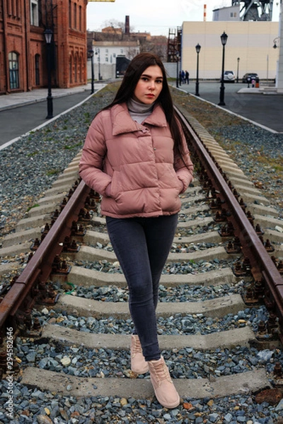 Obraz A young pretty pregnant girl in a pink warm down jacket walks near the old railway station. Late autumn, the cold is coming.