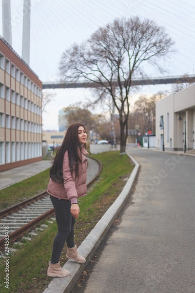 Obraz A young cute pregnant girl in a pink warm down jacket is trying to walk on a high curb. Late autumn, the cold is coming.