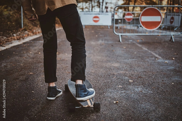 Fototapeta Young hipster is learning how to ride longboard while walking at autumn park.