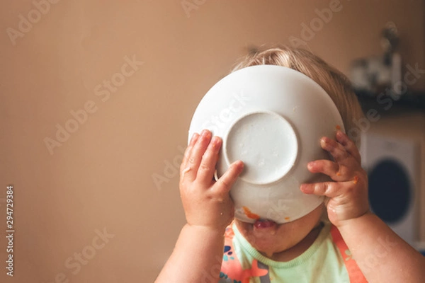 Obraz blond kid boy, very hungry eats and holds a plate with both hands, close-up with copyspace