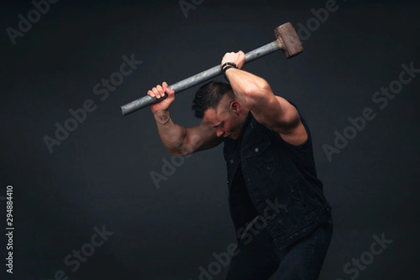 Obraz young bodybuilder in black clothes with a sledgehammer in his hands. Studio photography