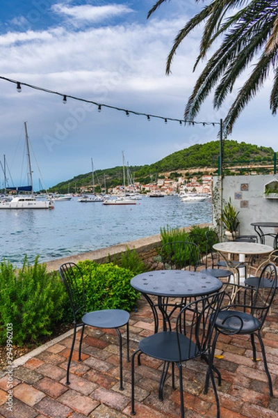 Fototapeta A beautiful Mediterranean beach bar terrace empty after rain decorated with flowers, palm trees and lights with a view of sailing boats in the port of Vis , Vis island, Croatia, Europe. Summer travel