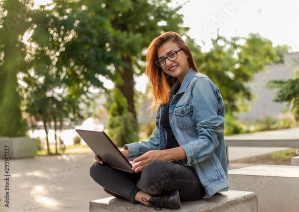 Fototapeta Young modern woman blogger in a denim jacket and glasses holds a laptop in his hands in the park. Online learning, blogging