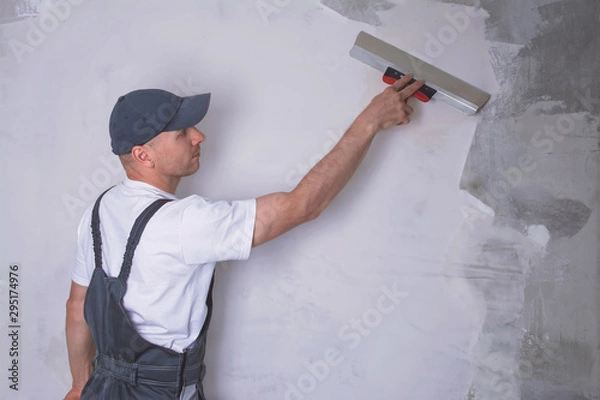 Fototapeta Worker in overalls plastering a wall with finishing putty using a putty knife. Repair work and construction concept
