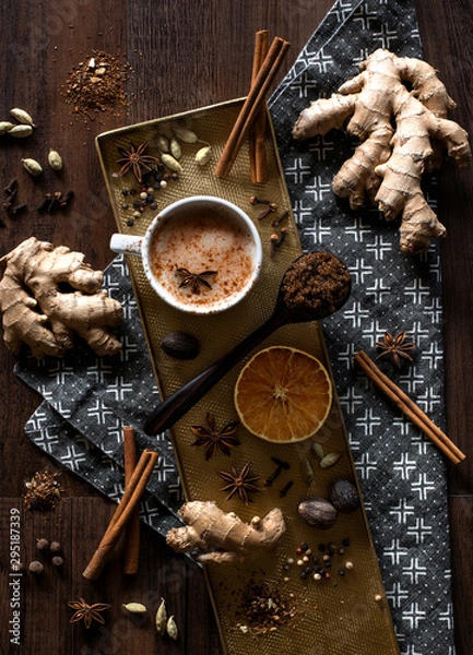 Obraz chai tea spices flat lay, chai tea latte with ginger root, cinnamon sticks, cardamom pods, star anise, whole nutmeg, peppercorns, whole cloves on a golden tray and grey patterned cloth napkin on table
