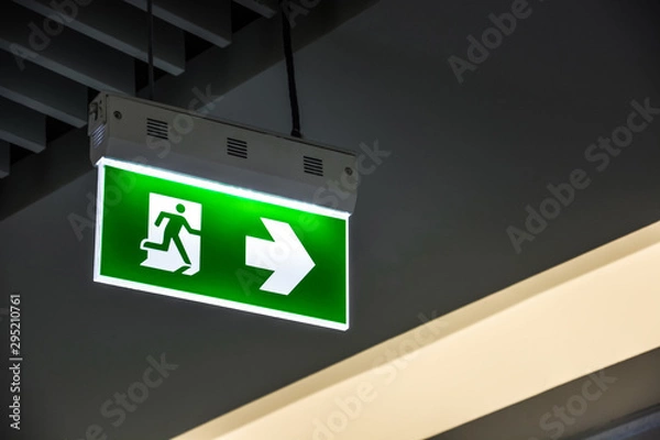 Fototapeta Illuminated green emergency exit sign hanging on ceiling in modern building