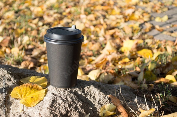 Fototapeta A black paper Cup of hot coffee stands on a stone, against a blurred background of autumn yellow leaves